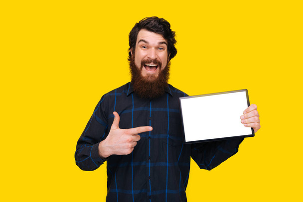 Φωτογραφία του ενθουσιασμένο άντρα με γενειάδα που δείχνει σε λευκή οθόνη του δισκίου, στέκεται πάνω από κίτρινο φόντο τοίχου  - Φωτογραφία, εικόνα