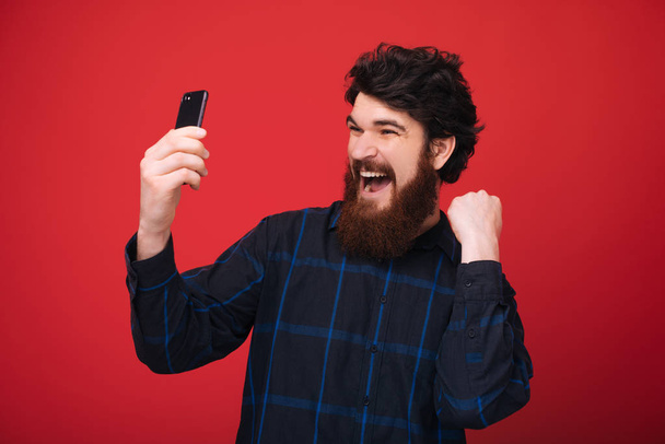 Захоплений бородатий хлопець тримає мобільний і святкує з піднятою рукою на червоному тлі
  - Фото, зображення