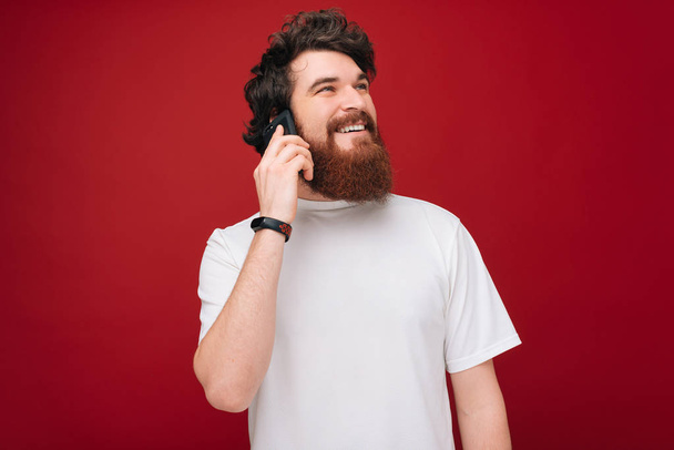 Vrolijke knappe bebaarde kerel in witte tshirt praten op telefoon over rode achtergrond  - Foto, afbeelding
