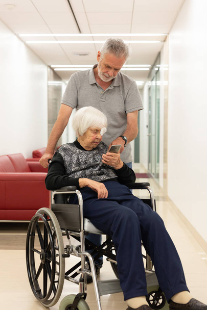 Чоловік середнього віку, який показує і допомагає літнім 95-річній жінці сидіти на інвалідному візку як користуватися сучасним мобільним телефоном
. - Фото, зображення