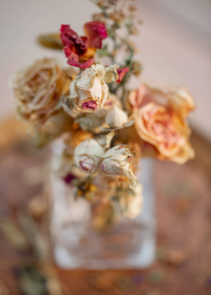 Καλλιτεχνική φωτογραφία. Τριαντάφυλλα μαραμένα σε ένα γυάλινο βάζο. Ξεθωριασμένα τριαντάφυλλα και ξηρό γρασίδι σε μια ξύλινη επιφάνεια. Φύση ταπετσαρία θολή φόντο. Ήπια εικόνα. Closeup. Απαλή εστίαση. - Φωτογραφία, εικόνα