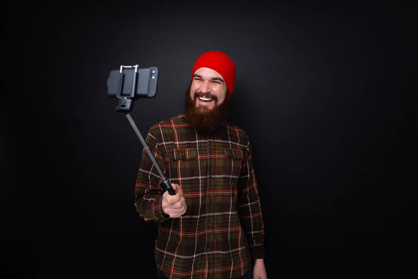 Sakallı mutlu bir adamın karanlık bir arka plan üzerinde izole sopaile selfie fotoğrafı yapma portresi  - Fotoğraf, Görsel