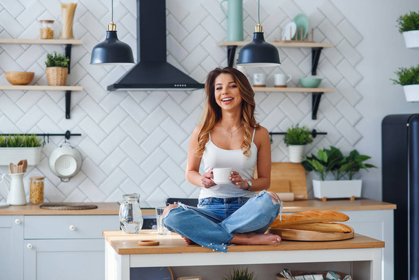 Mujer bastante feliz sentado en la mesa en la acogedora cocina por la mañana en casa y bebe café. Relajación matutina en casa
. - Foto, imagen