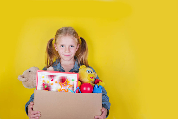 blondes kleines Mädchen mit einer Schachtel mit Spielzeug auf gelbem Hintergrund. Spendenkonzept.. - Foto, Bild