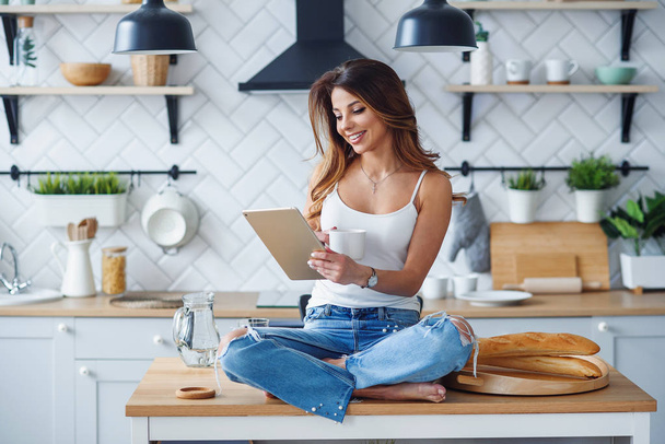 Beautiful woman sitting on the table and uses tablet computer on the cozy kitchen in the morning at home. - Foto, immagini