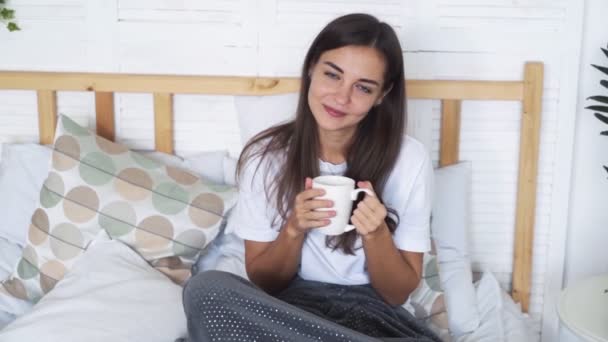Beautiful girl sits on bed, holds cup, looks at camera and smiles, slow motion - Πλάνα, βίντεο