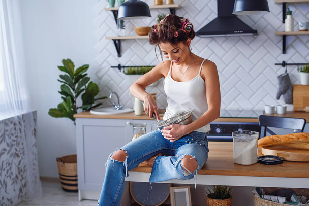 Beautiful smiling woman in casual clothes kneads a dough in a metal plate at stylish kitchen. Young pretty woman baking on bright home kitchen. - Photo, image