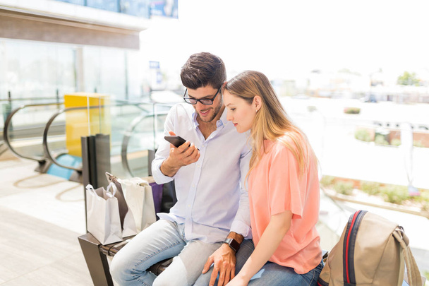 Jeune couple vérifiant les offres sur smartphone tout en étant assis dans le centre commercial
 - Photo, image