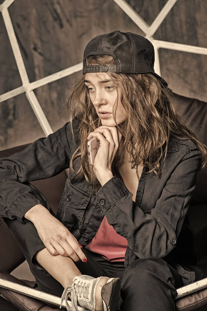 Beauty girl with hipster look and style. Sensual woman in cap on long hair. Woman with thinking face relax in armchair. New technology for modern life. Fashion and beauty look, vintage filter - Fotó, kép