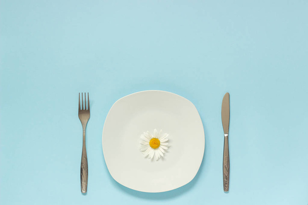 One flower chamomile daisy on plate, cutlery fork knife on blue paper background Concept vegetarian, healthy eating, diet or anorexia Creative top view Copy space template for lettering text or design - Фото, зображення