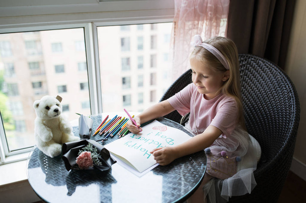 Child daughter painting postcard for mom. Girl sitting at home on table, next lying pink flower for mother. Mother day concept - Фото, изображение