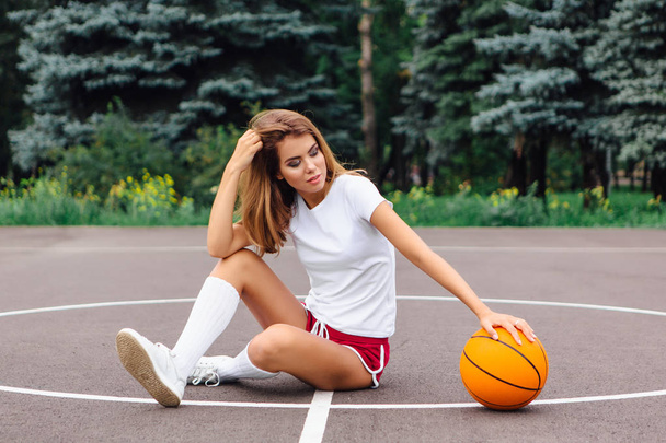 Krásná mladá dívka oblečená do bílých triček, kalhot a tenisky, sedí na basketbalovém hřišti s koulí. - Fotografie, Obrázek