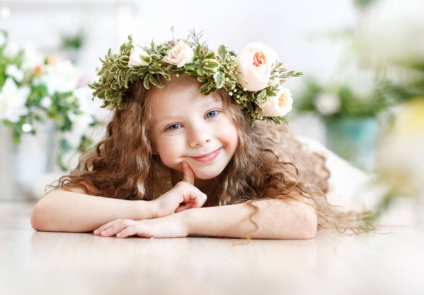 Kid - girl with blond hair in a flower crown. Beautiful little girl in a bright studio lying on the floor and looks into the camera - Фото, зображення