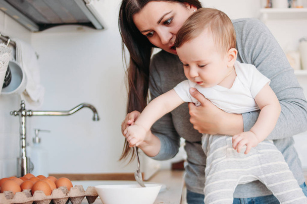 Young mother brunette woman with baby boy in arms cooking food in white modern kitchen at home - 写真・画像