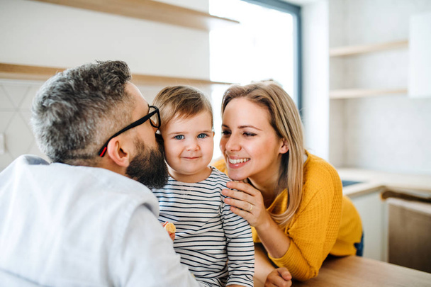 A portrait of young family with a toddler girl indoors in kitchen, kissing. - Foto, Bild