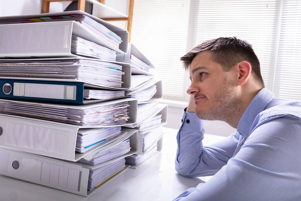 Rear View Of Stressed Businessman With Hand On Head In Front Of Stacked Folders - Photo, Image