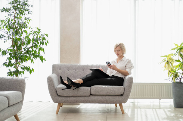 Young beautiful blonde girl with short hair in a white shirt is sitting on the sofa in bright in the office against the window. Holds a notebook and smartphone. Talking on the phone. - Photo, Image