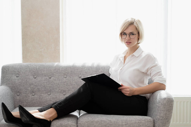 Young beautiful blonde girl with short hair in a white shirt is sitting on the sofa in bright in the office against the window. Holds a notebook and smartphone. Talking on the phone. - Фото, изображение