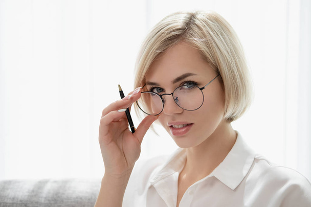Young beautiful blonde girl with short hair in a white shirt and glasses close-up holds a pen in her hands. Holds glasses with his hands and looks out from under eyeglasses. - Fotoğraf, Görsel
