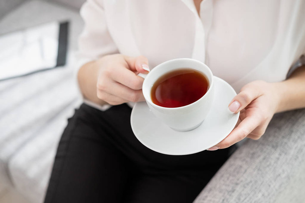Young beautiful blonde girl with short hair in a white shirt and glasses is sitting on the sofa in bright in the office against the window. Holds a white cup and saucer. Drinking coffee or tea in a - Фото, изображение