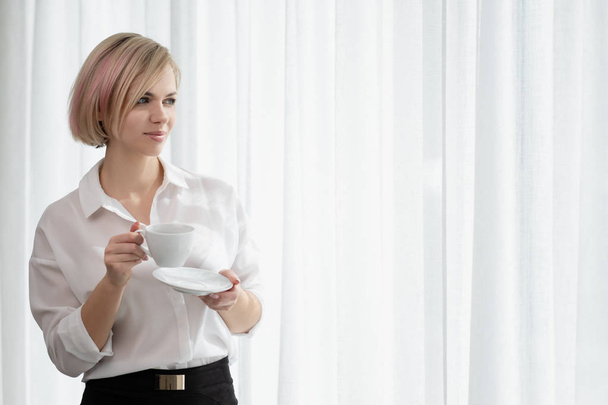 Young beautiful blonde girl with short hair in a white shirt and glasses is sitting on the sofa in bright in the office against the window. Holds a white cup and saucer. Drinking coffee or tea in a - Valokuva, kuva