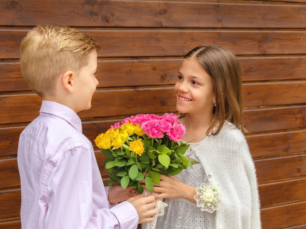 Cute little boy giving bouquet of flowers to charming little lady, smiling girl in love receiving yellow and pink roses from friend, generation z friendship concept with two happy kids outdoor - Photo, image