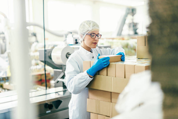 Молода жінка-працівник у стерильній уніформі підраховує коробки, стоячи на харчовій фабриці
. - Фото, зображення
