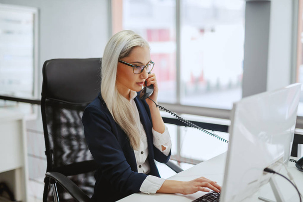 Jeune femme d'affaires blonde caucasienne en tenue formelle et avec des lunettes tapant sur le clavier et ayant un appel téléphonique alors qu'elle est assise au bureau. Chaque jour est un nouveau jour. Traitez-le ainsi.
. - Photo, image