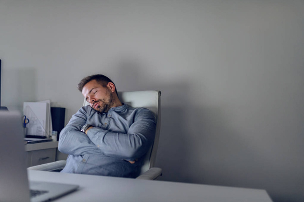 Tired handsome Caucasian bearded employee sleeping in the chair late at night in home office. - Photo, Image