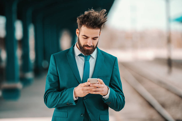 handsome Caucasian bearded businessman in turquoise suit using smart phone while waiting for train to arrive. Opportunities don't just happen, you create them. - Foto, imagen