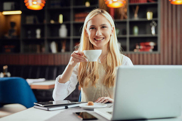 Charmante lachende blonde zakenvrouw gekleed Smart Casual zitten in cafetaria, koffie drinken en laptop gebruiken. - Foto, afbeelding
