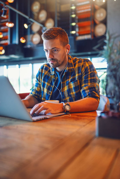 Fiatal jóképű fehér blogger kockás ingben és fülhallgatóval a fülében laptoppal, miközben a kávézóban ül. - Fotó, kép