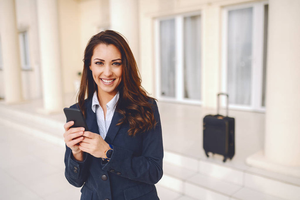 Young smiling Caucasian businesswoman dressed smart casual using smart phone for writing message while standing outdoors. In background luggage. Business trip concept. - Φωτογραφία, εικόνα