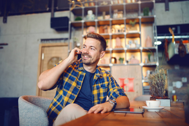 Young Caucasian handsome unshaven man in plaid shirt sitting in cafe and talking on the phone. On desk tablet and coffee. - Photo, image