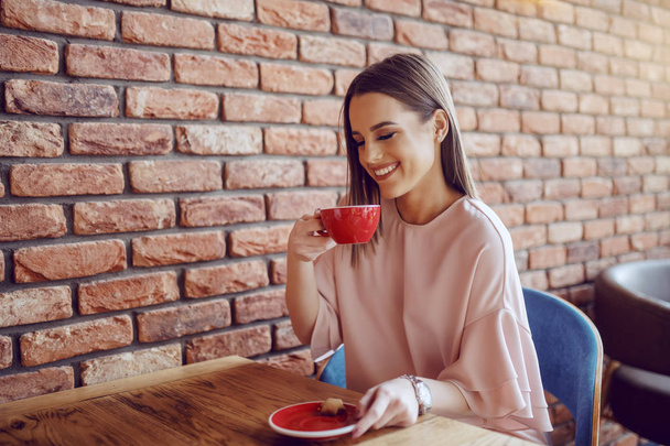 Smiling Caucasian brunette with eyes closed dressed elegant enjoying coffee in cafeteria. - Photo, Image