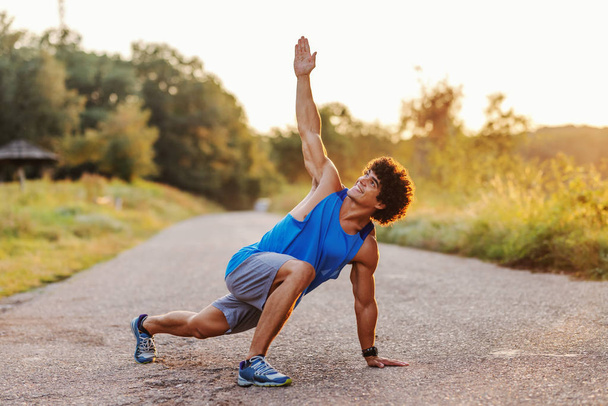 Smiling strong Caucasian man in sportswear and with curly dark hair doing fitness exercises on the country road on sunny summer day. Fitness in nature concept. - Photo, Image
