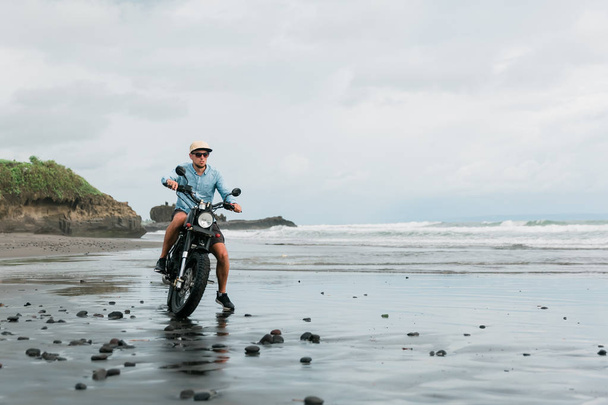 Man in cap riding motorcycle on beach. Moto cross dirtbiker on beach sunset on Bali. Young hipster male enjoying freedom and active lifestyle, having fun on a bikers tour. - Photo, Image