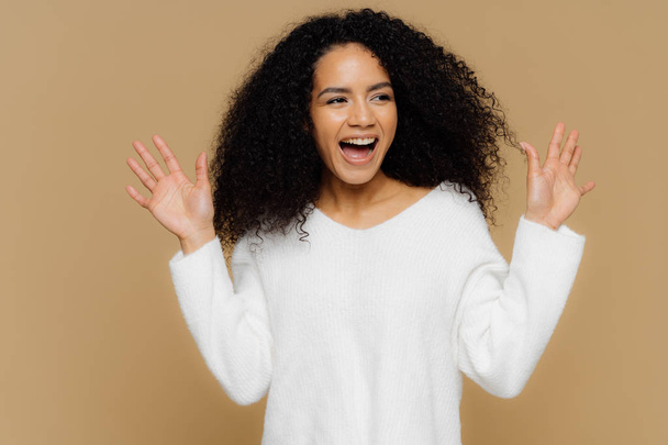 Glad overemotive woman surprised by pleasant relevation, raises hands and shows palms, focused aside, wears casual white sweater, models over brown background. Positive human emotions and feelings - Photo, image