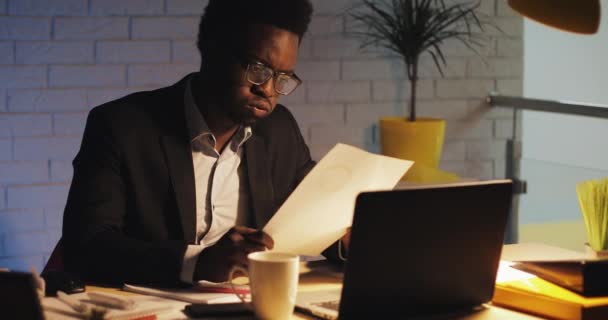 Tired young black businessman with laptop computer and papers working at night office. He working with documents. Business, workaholic, deadline concept. - Záběry, video