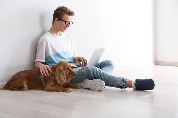 Adolescente chico con lindo perro y portátil sentado cerca de la pared de luz
 - Foto, imagen