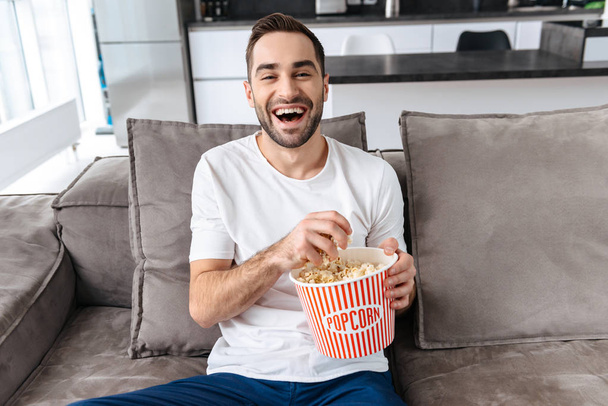 Photo of unshaved man 30s in casual t-shirt holding popcorn bucket and remote control while sitting on couch in living room - Valokuva, kuva