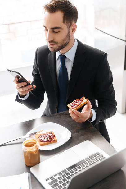 Image of brunette businessman having breakfast while using smart - Zdjęcie, obraz