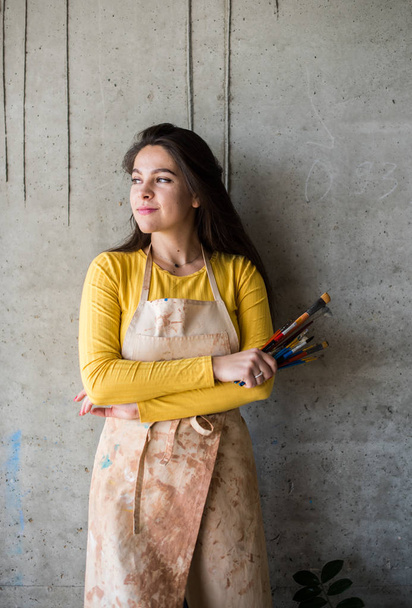 Jonge mooie dame artiest in schort met verf vlekken in haar loft artistieke studio - Foto, afbeelding