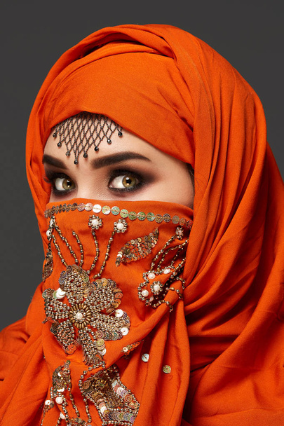 Studio shot of a young charming woman wearing the terracotta hijab decorated with sequins and jewelry. Arabic style. - Foto, afbeelding