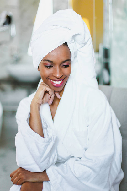 Young cute afro american girl sitting in bathroom and wearing white bathrobe. - 写真・画像