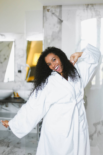 Young afro american smiling woman dancing in bathroom and wearing white bathrobe. - Fotoğraf, Görsel