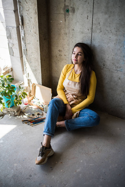 Nuori kaunis nainen taiteilija esiliina maalitahroja istuu lattialla hänen studio
 - Valokuva, kuva