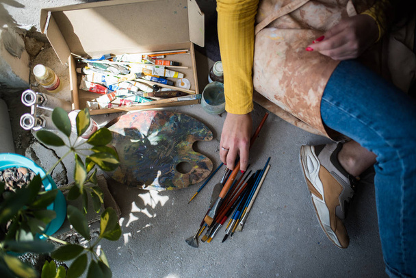 Młoda piękna artystka w fartuchu z plamami po farbie siedząca na podłodze w swoim studio - Zdjęcie, obraz