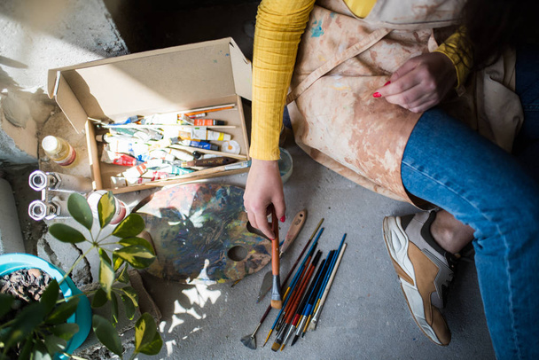 junge schöne Künstlerin in Schürze mit Farbflecken auf dem Boden in ihrem Atelier - Foto, Bild