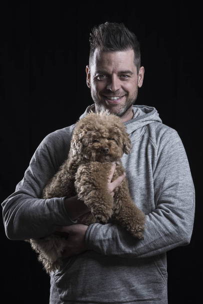 Portrait of a smiling man looking at the camera with a red poodle dog. Black background. Vertical. - Фото, изображение